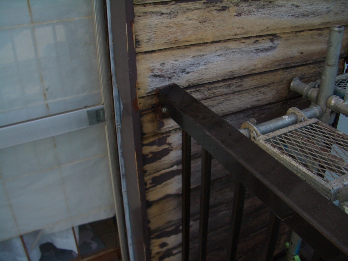 Close-up of a weathered wooden exterior wall with peeling paint, a dark metal railing extending from the foreground, and a rusted metal grate platform with pipes on the right.