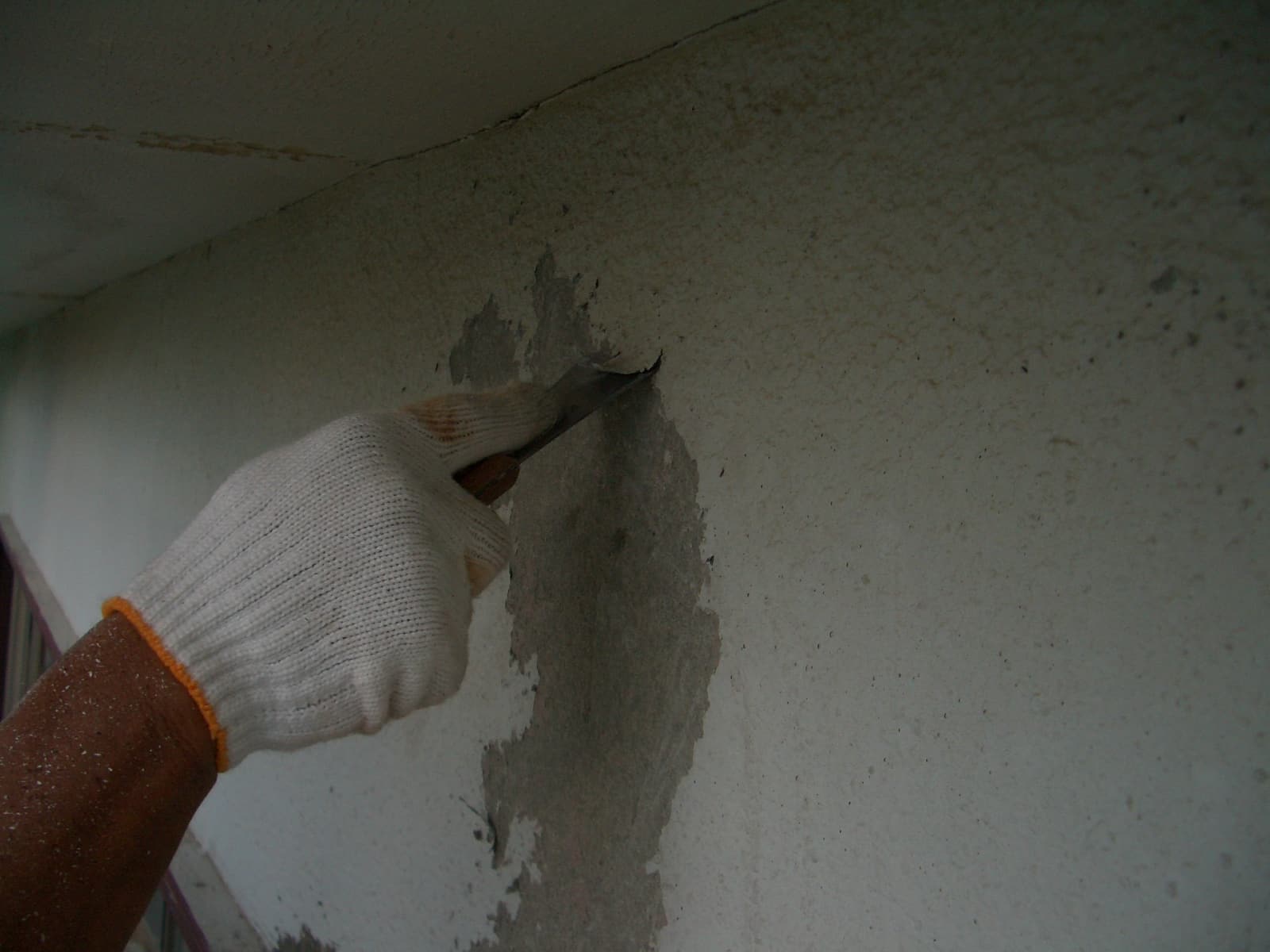Gloved hand scraping peeling plaster off a wall with a putty knife.