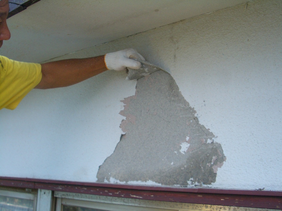 Worker in a yellow shirt and white glove peeling plaster from an exterior wall, revealing a large gray patch of concrete surface