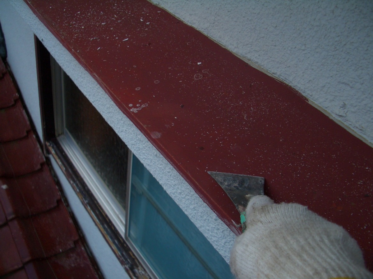 Person wearing a white glove uses a scraper to remove debris from a red metal window flashing on a sloped roof edge.
