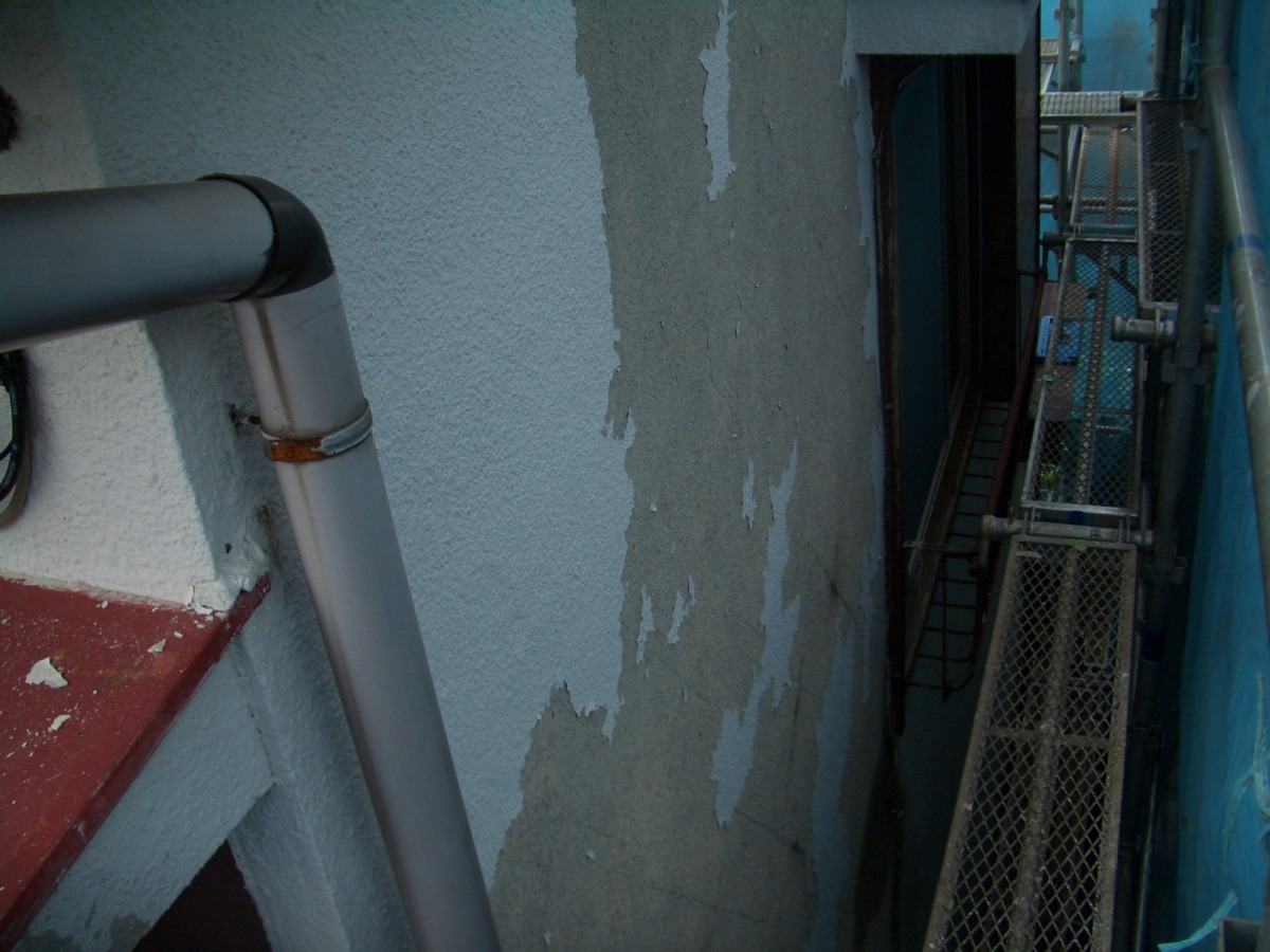 Exterior wall with peeling paint beside a gray downspout pipe and metal scaffolding on the right side of the image