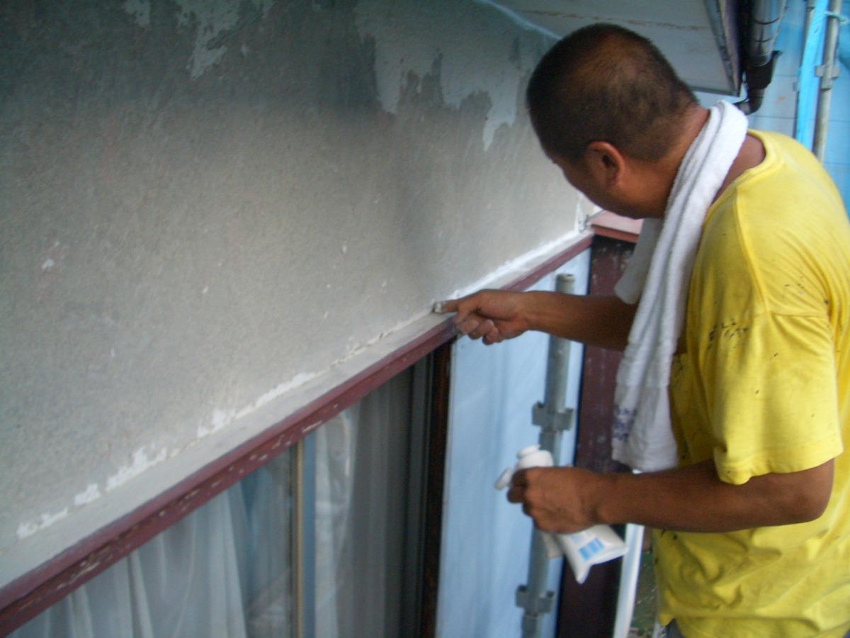 Painter on scaffolding applying paint to a window frame on a building, wearing a yellow shirt and a white towel draped around his neck.