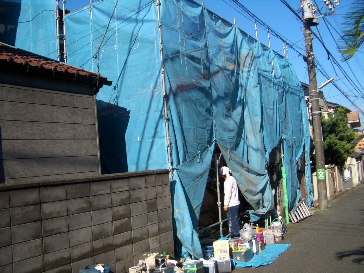 Construction site along a street with scaffolding covered in blue tarps; a worker in white helmet stands among tools and buckets.