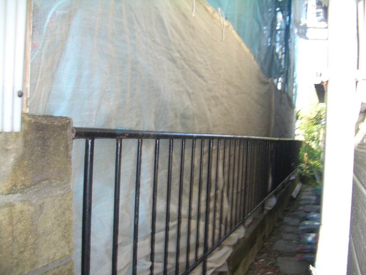 Narrow outdoor alley with a black metal railing beside a concrete wall covered by a blue tarp.