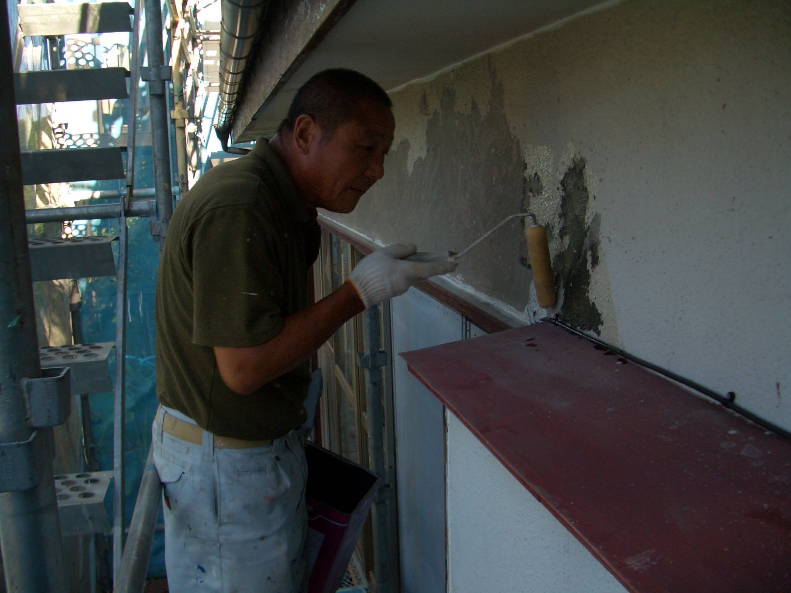 A man on scaffolding painting a wall with a roller using white/gray paint on an exterior surface.