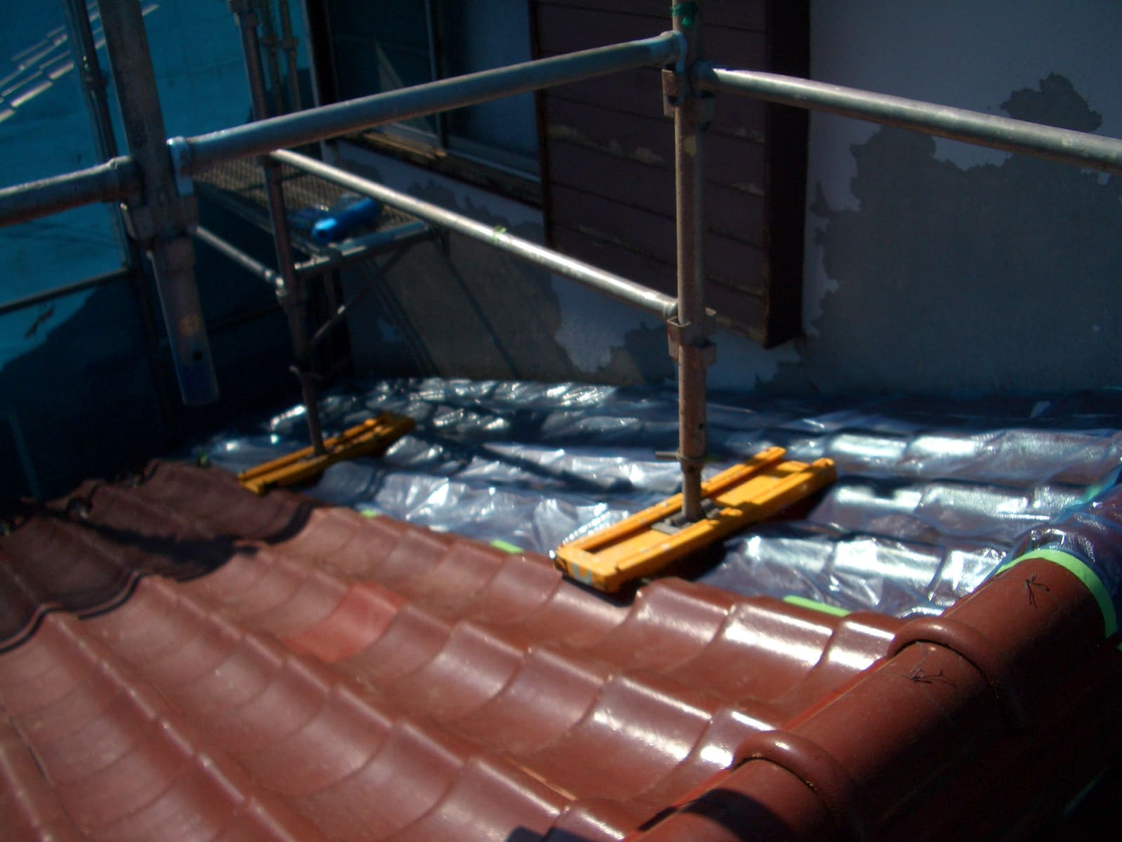 Scaffolding on a roof with tile shingles and a blue underlayment, orange spacers resting on the membrane