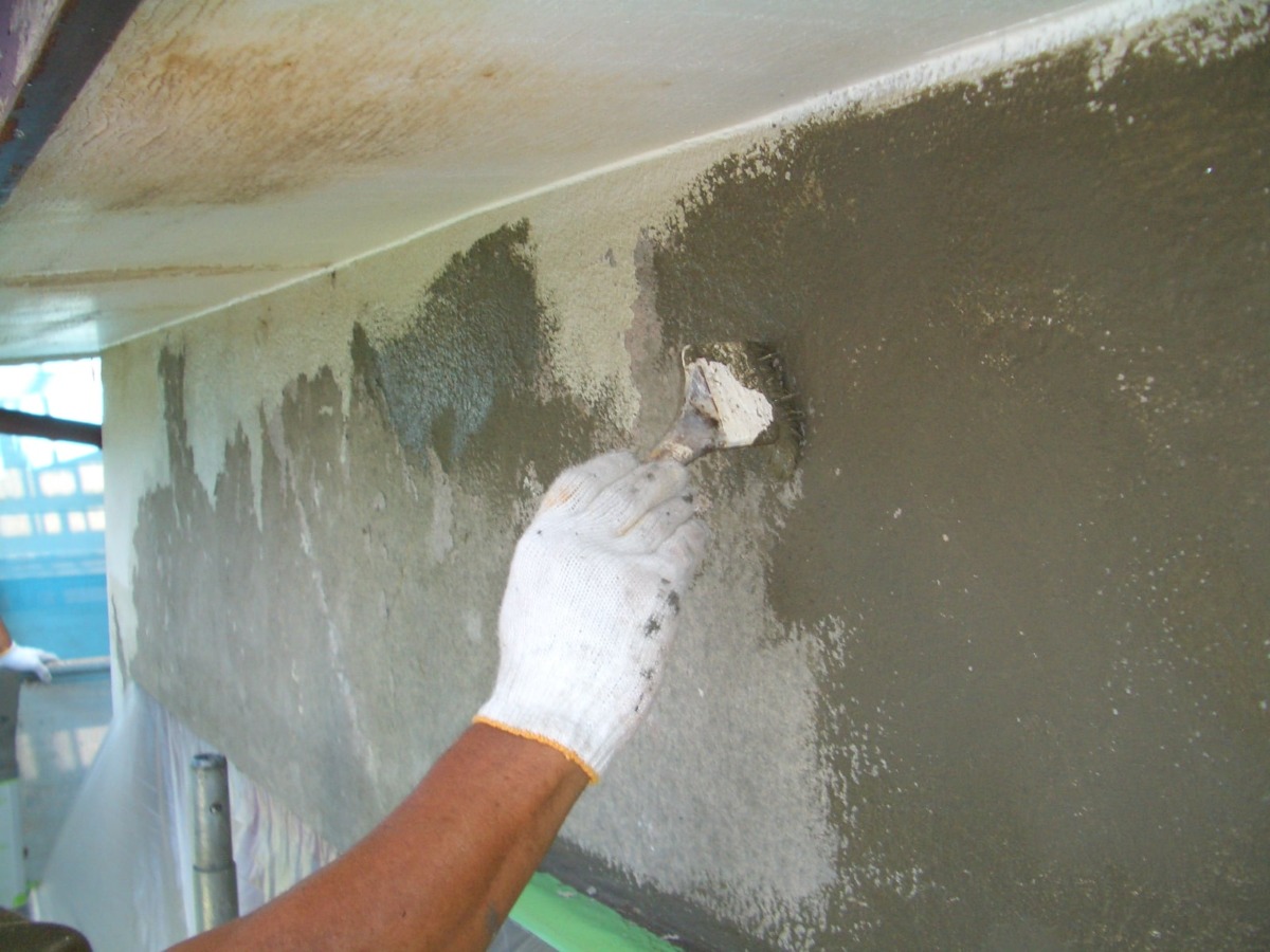 Worker's gloved hand smoothing gray plaster onto a wall with a trowel under an overhang