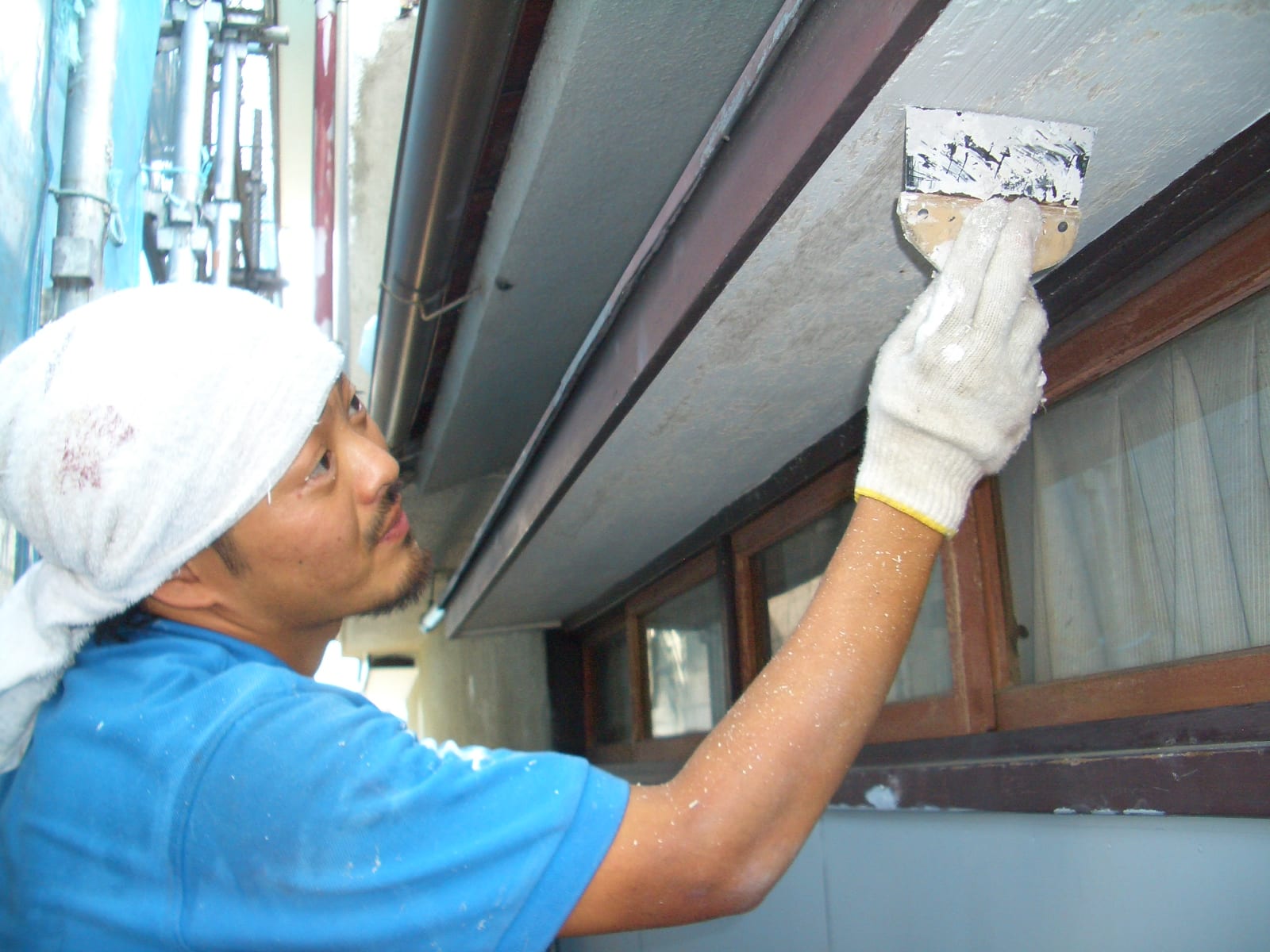 Man painting an exterior wall with a wide brush, wearing a white head wrap and gloves, blue shirt visible in a narrow alley.
