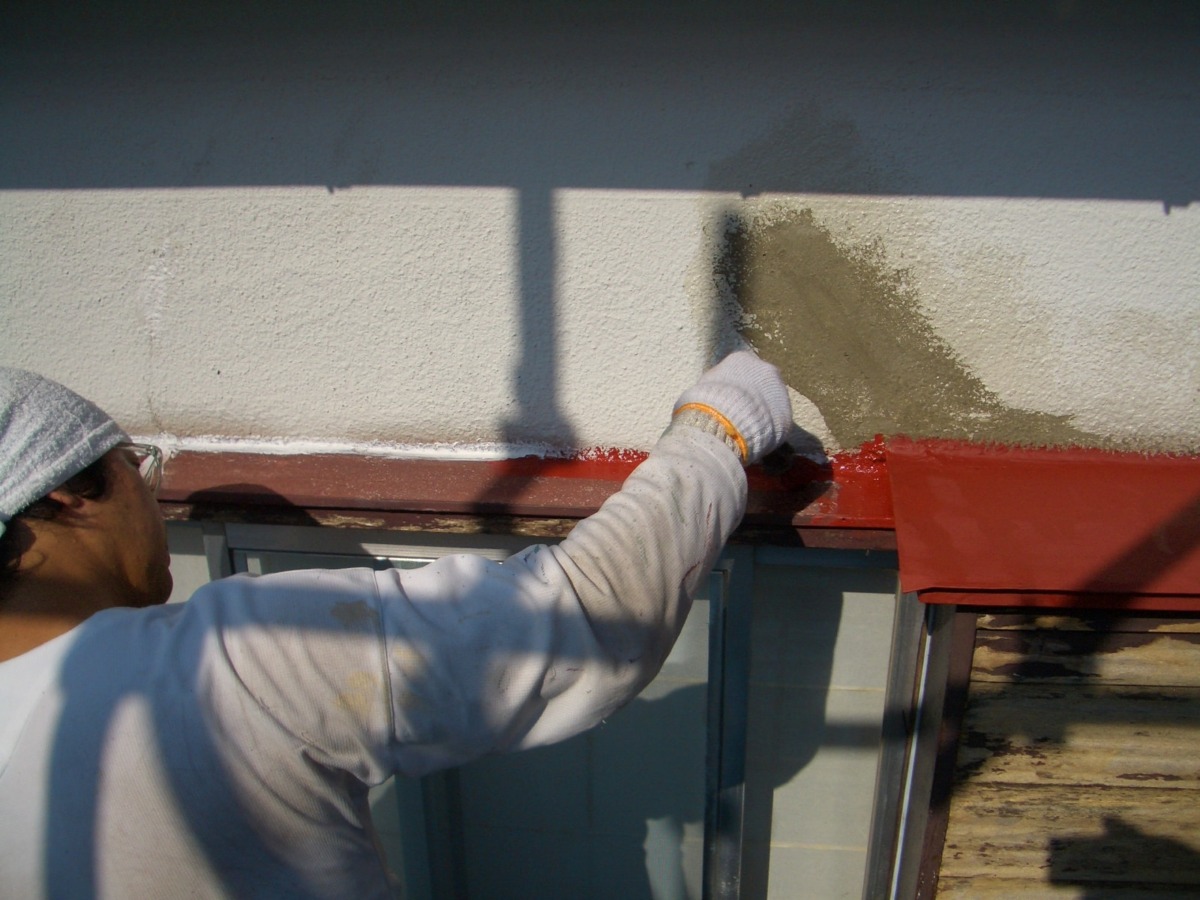 Worker sealing exterior wall above a window with a brush, wearing a white glove and light-colored sweater.