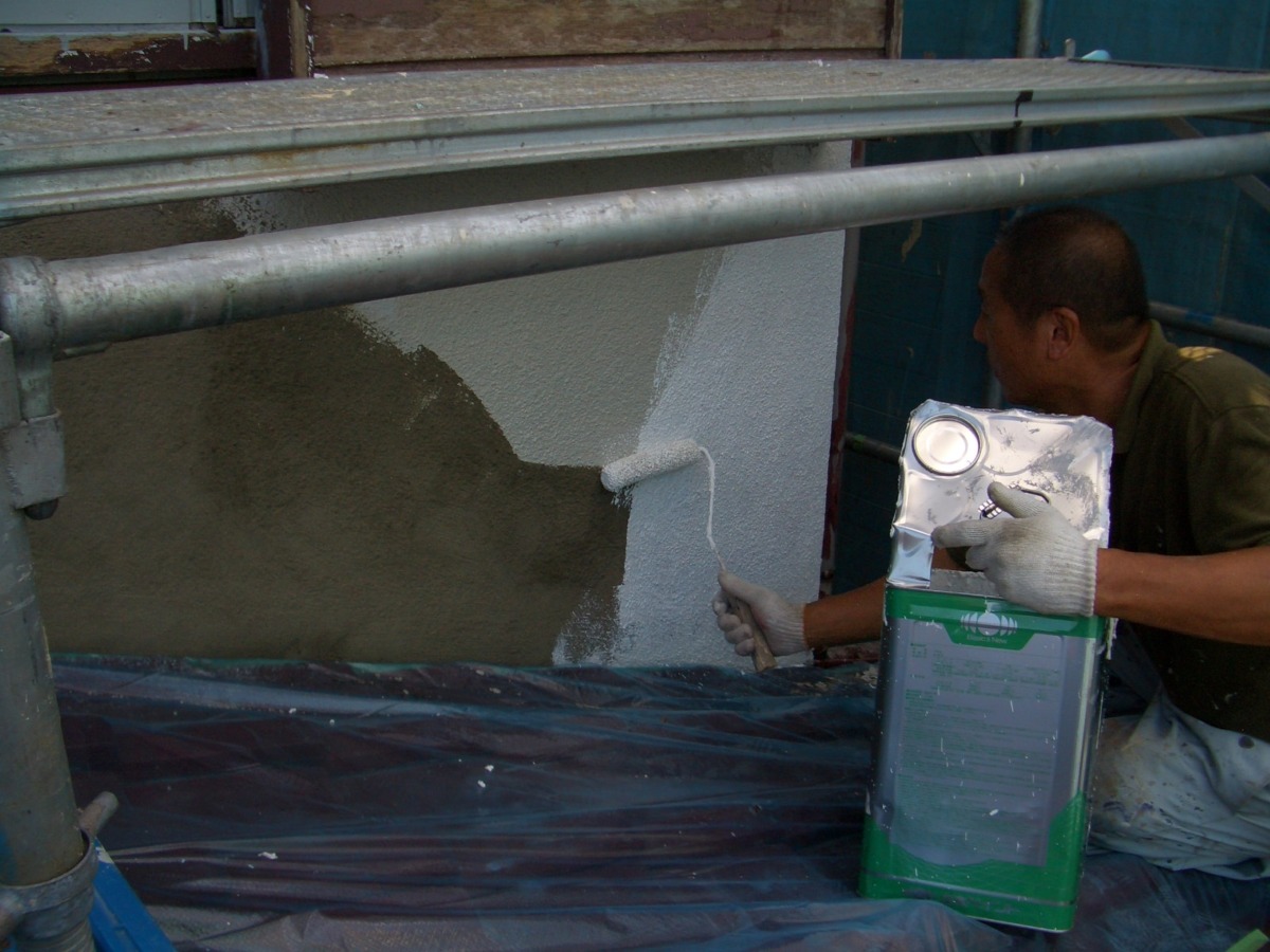 Worker using a paint roller to apply gray plaster to an exterior wall, holding a metal paint bucket beside him under scaffolding