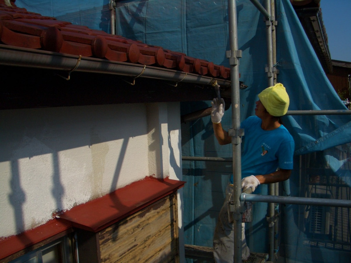 Construction worker on scaffolding painting or repairing eaves of a building, wearing a blue shirt and yellow head covering, gloves, and reaching to the roof edge with a tool.