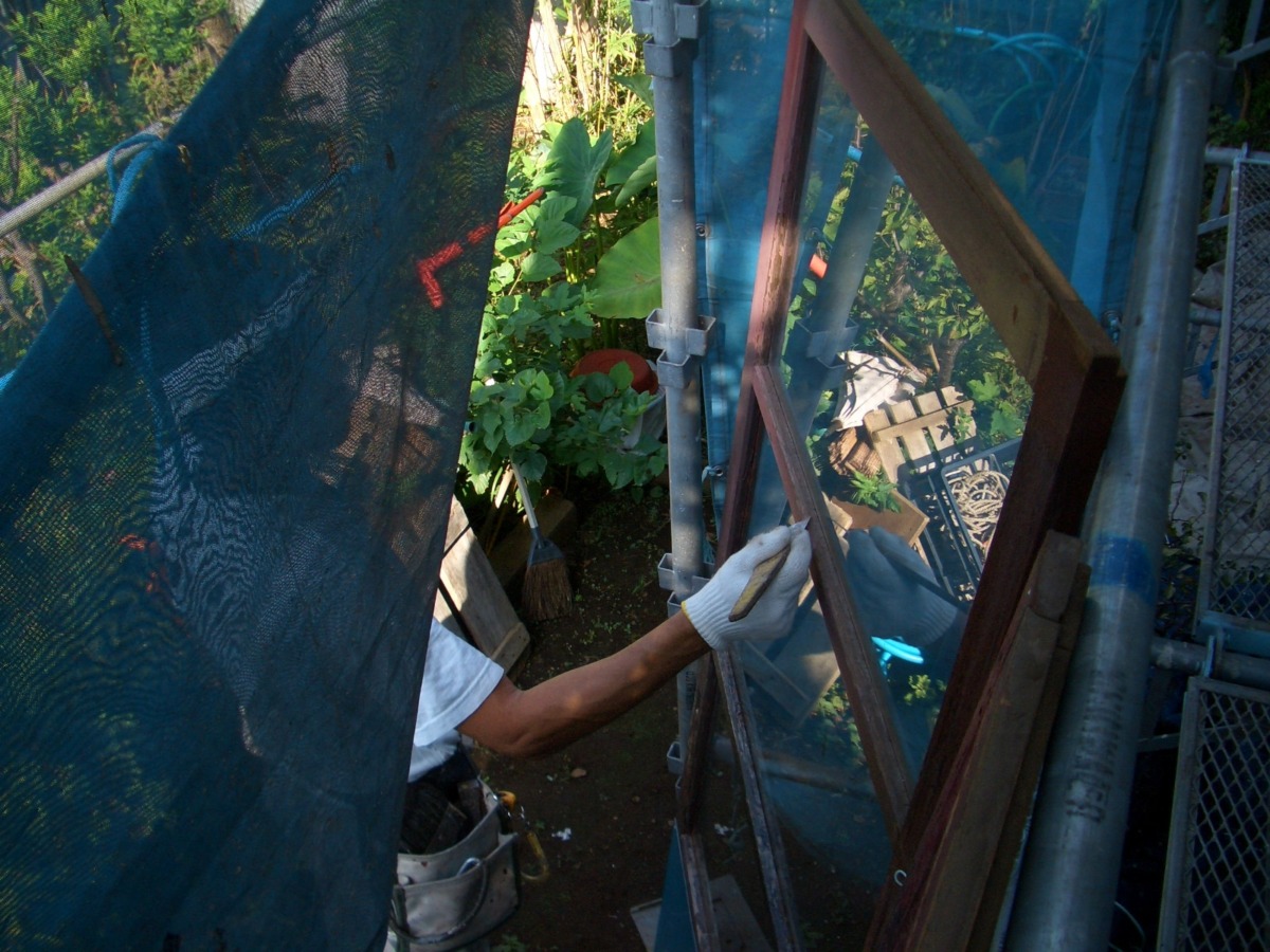 Worker wearing white gloves painting a wooden window frame on a blue scaffold, outdoors on a sunny day
