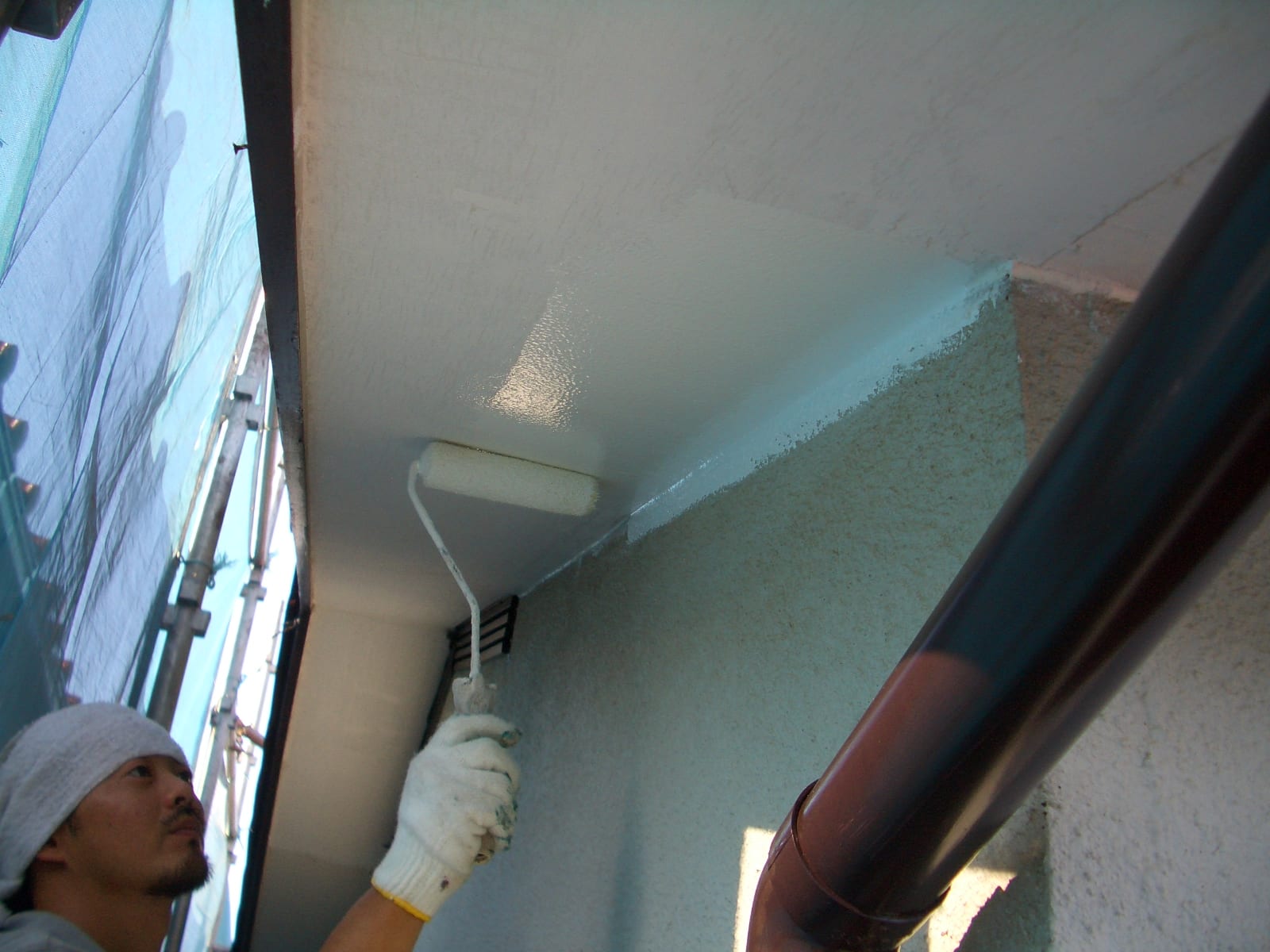 Worker wearing a gray beanie and glove rolling light blue paint onto a ceiling with a paint roller in a narrow outdoor area.