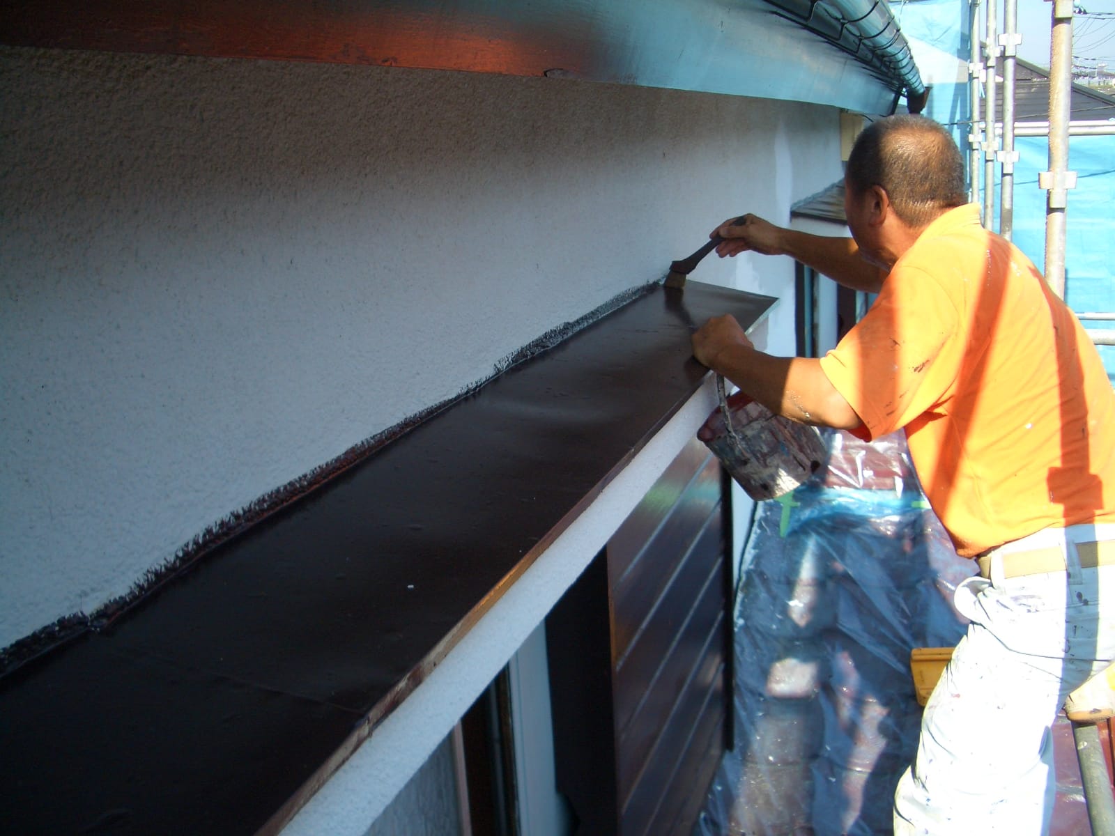 Construction worker in orange shirt painting a windowsill with a brush along an exterior wall.