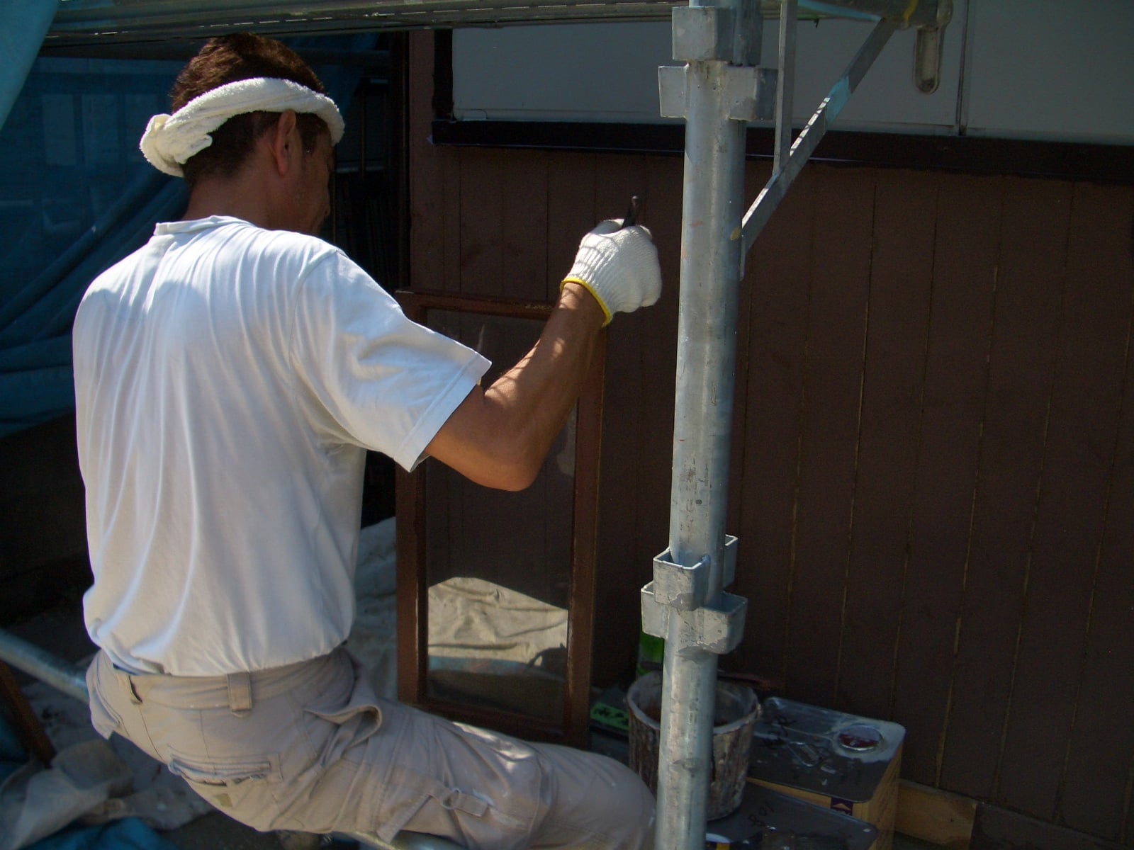 Construction worker wearing a white shirt and headband, kneeling on scaffolding while painting a wooden fence or wall brown with a brush/glove on.