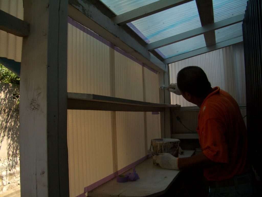 Worker in an orange shirt paints a wooden frame inside a shed-like structure, a paint bucket nearby.
