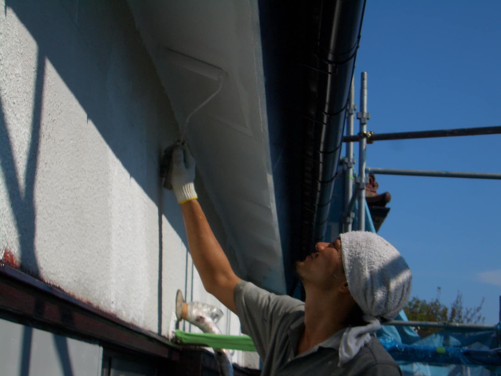 A worker in a glove paints the exterior wall using a roller while standing on scaffolding outdoors, with a cloth wrapped around their head.