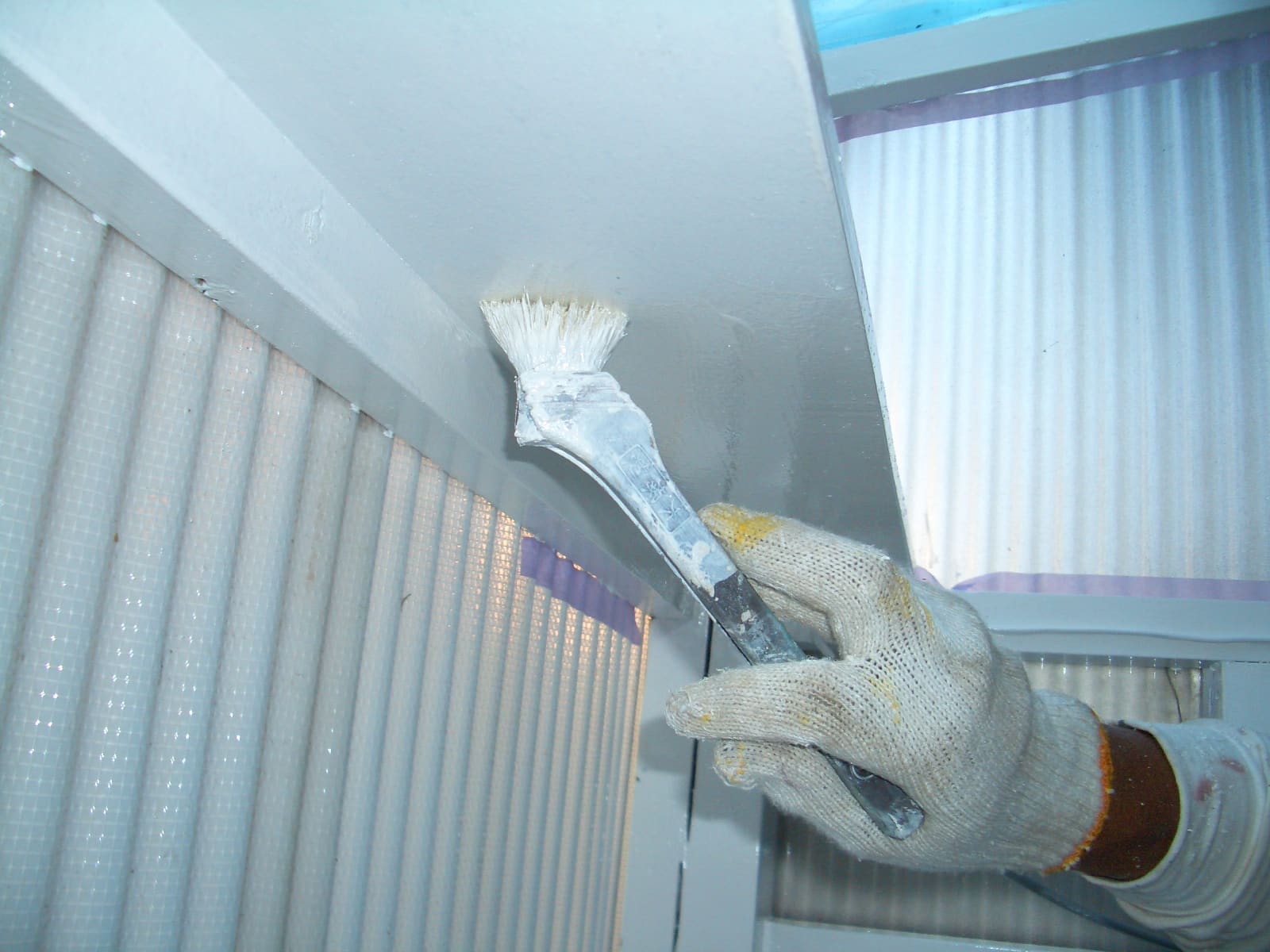 Gloved hand painting a ceiling with a flat brush indoors near a windowed wall.