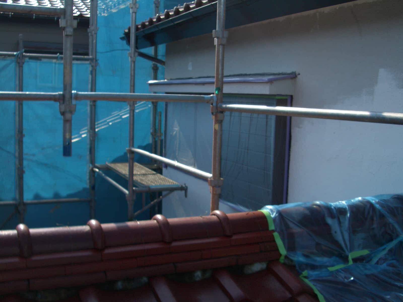 Construction scaffolding with metal poles surrounding a building under renovation, blue tarp in the background and neatly stacked red roof tiles in the foreground.