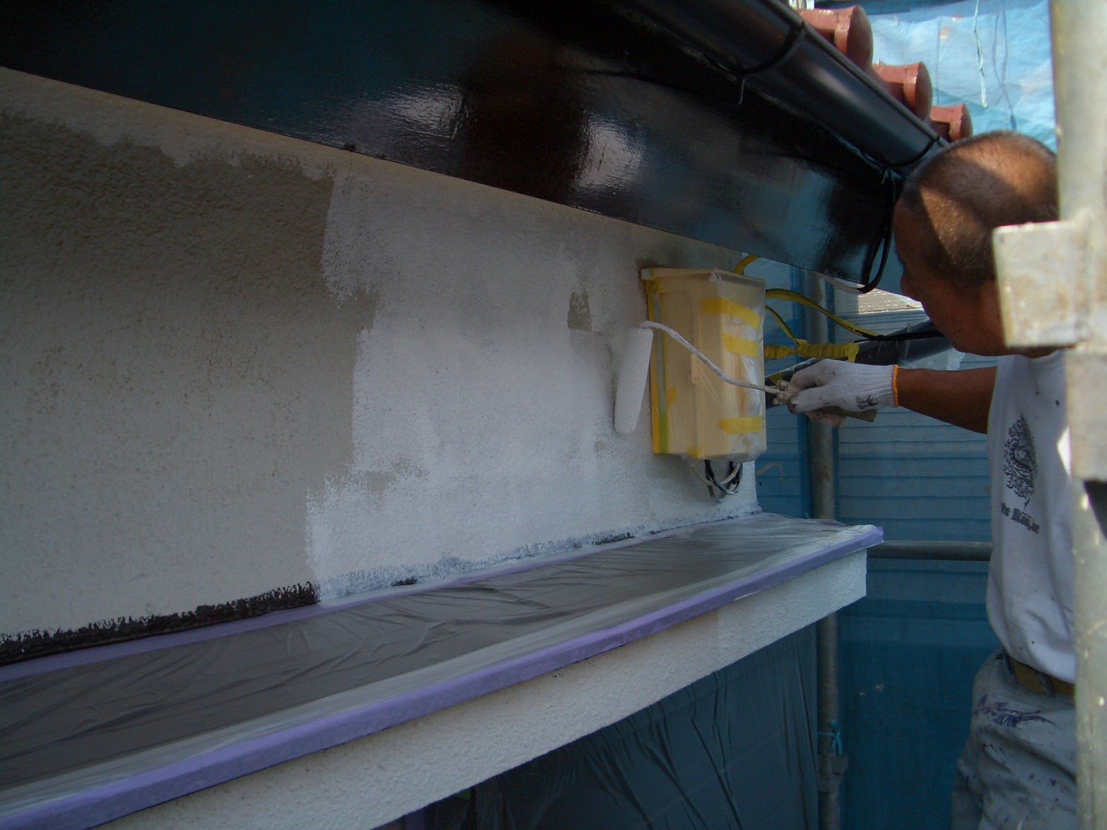 Painter rolling white paint onto an exterior wall next to a taped electrical box, with scaffolding and blue tarps in the background.