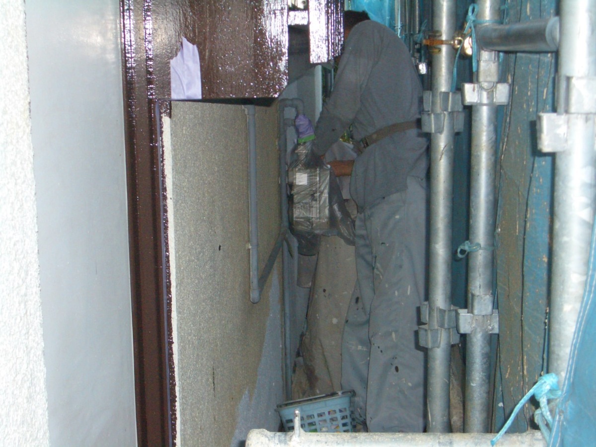 Worker in gray coveralls and gloves works in a narrow utility alley with exposed pipes and scaffolding, handling a bag or bundle near a wall.