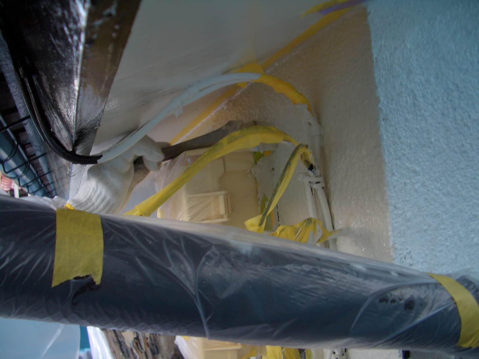 View of a construction site ceiling with insulated pipes, yellow tape, and plastic sheeting covering surfaces.
