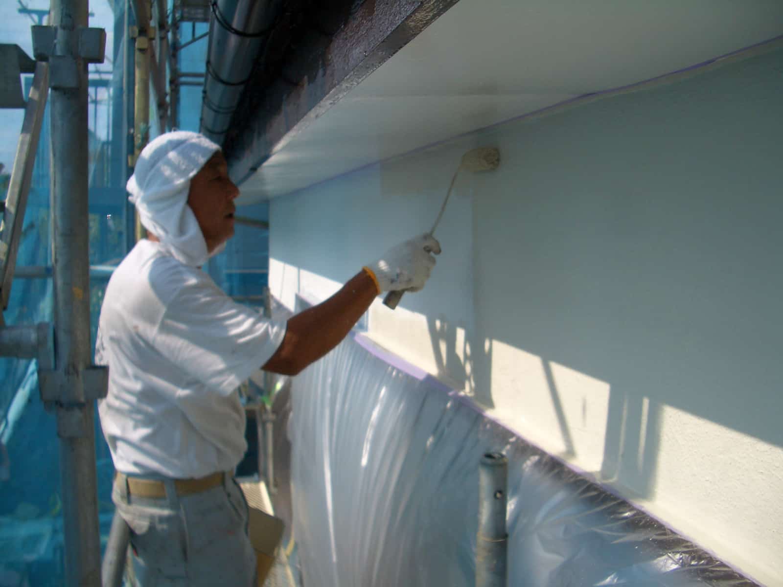 A construction worker in white protective clothing painting a wall with a roller while standing on scaffolding.