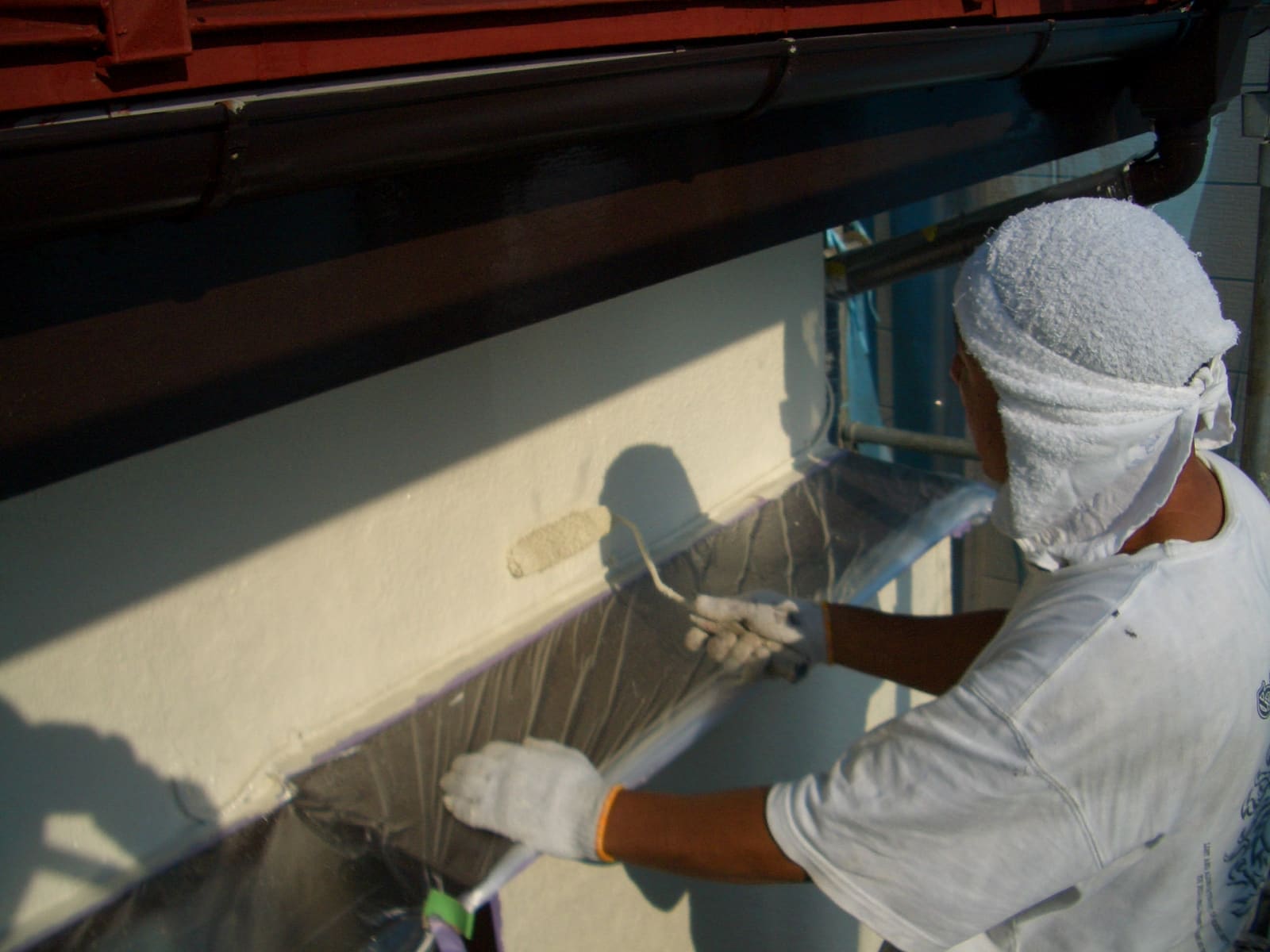 Worker in white clothing and a head wrap paints an exterior wall with a roller on scaffolding.