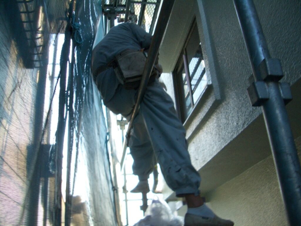 Construction worker in gray coveralls climbing a scaffold beside a building, performing exterior maintenance.