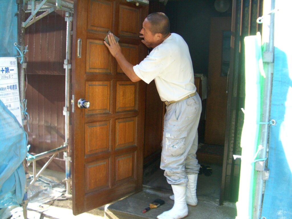 Construction worker in white shirt and boots opens a large paneled wooden door at a building under renovation.