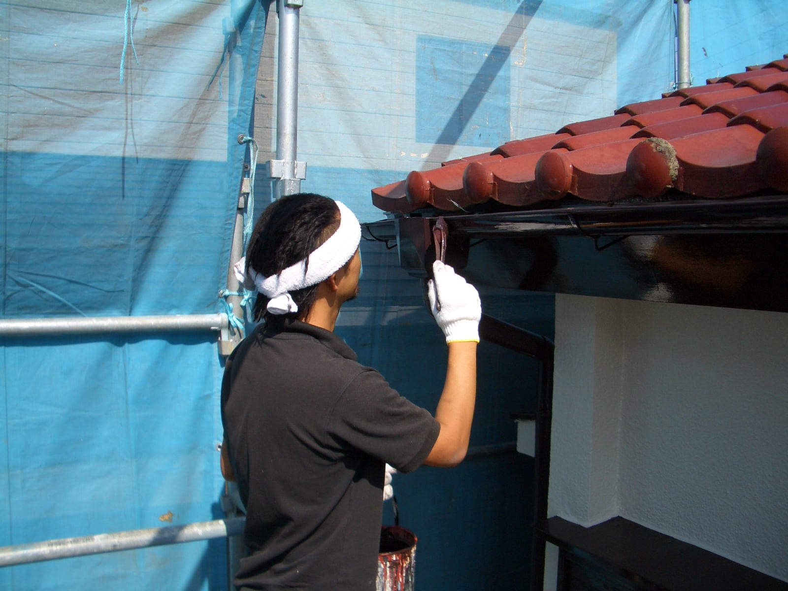 Worker with white headband and gloves painting or sealing a roof edge under red clay tiles at a construction site with blue scaffolding netting nearby