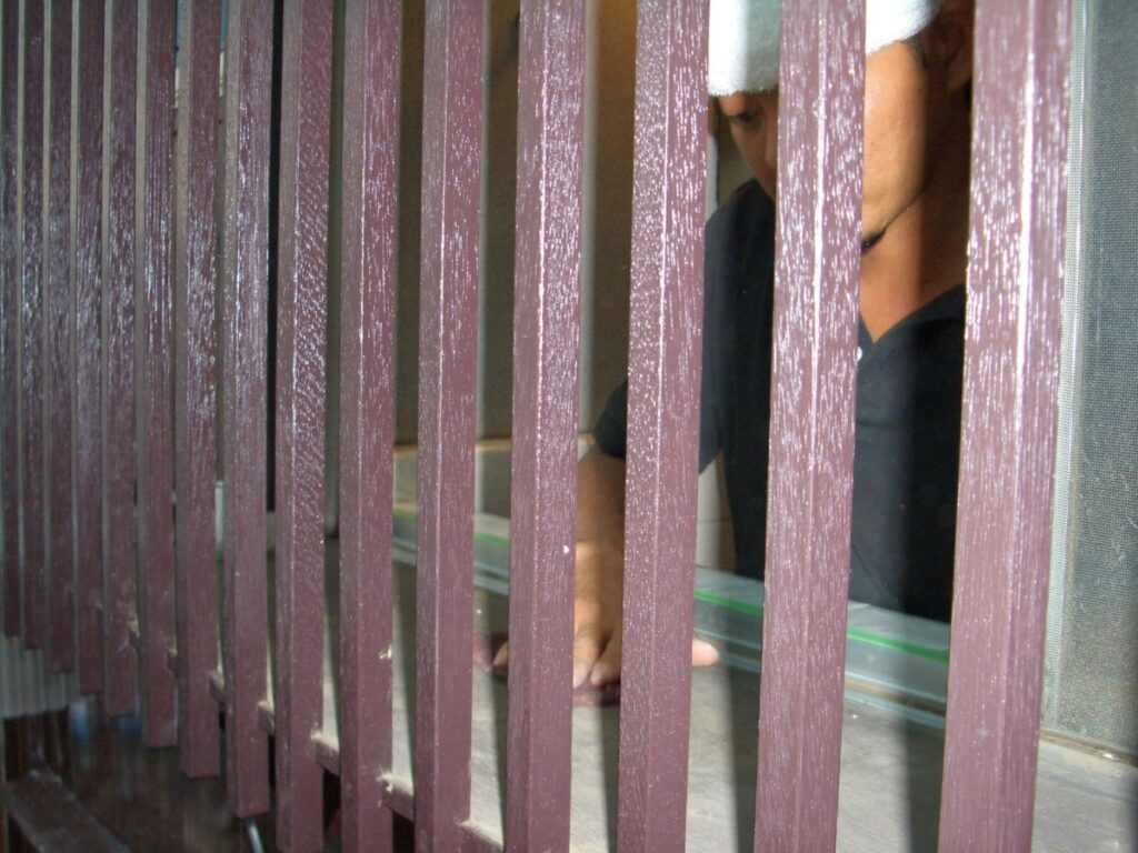 Person behind purple vertical bars at a counter, reaching with a hand toward a glass surface, partial face visible through the gaps.