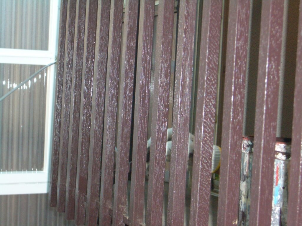 Close-up of a brown wooden vertical railing or fence with a building window visible on the left side in the background.