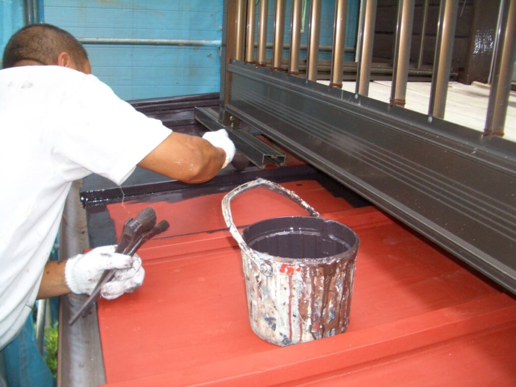 Worker in a white shirt and gloves paints a red metal roof, with a paint bucket and brushes nearby.