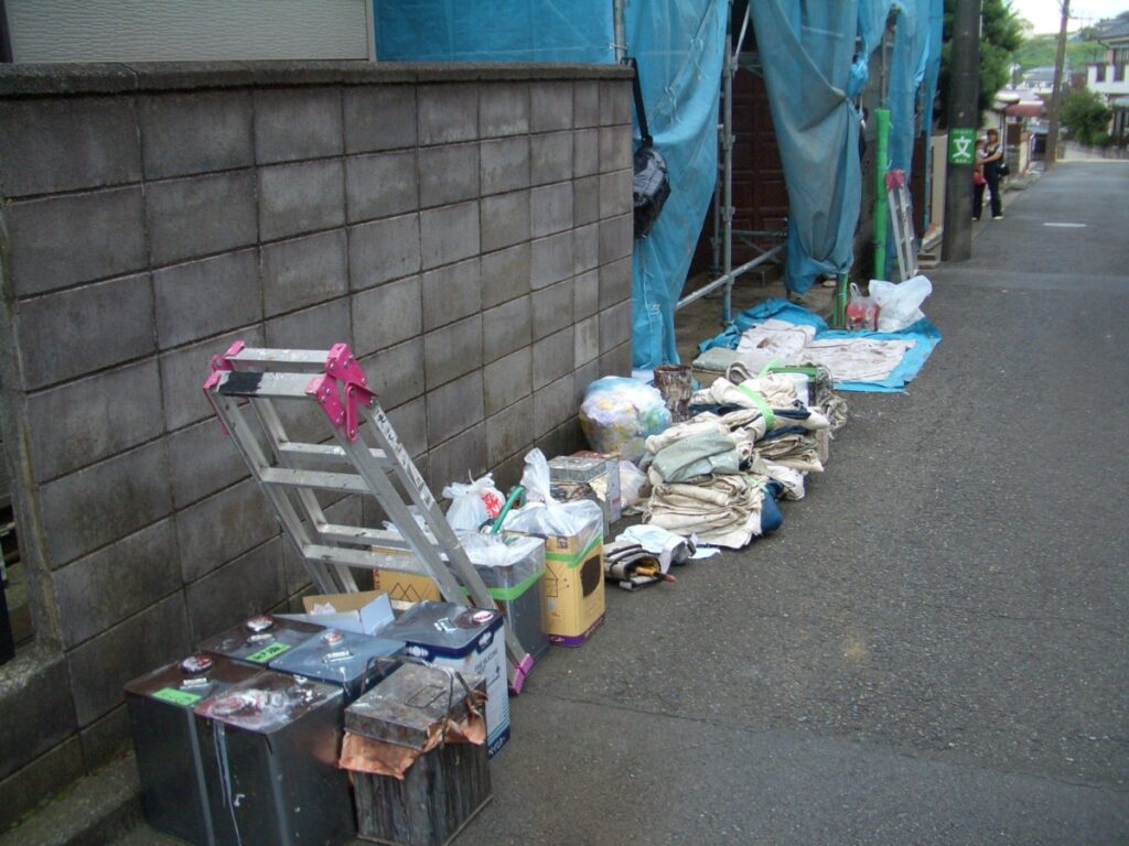 Construction site trash lined along a sidewalk with blue tarps, scaffolding, ladders, and bags of debris.