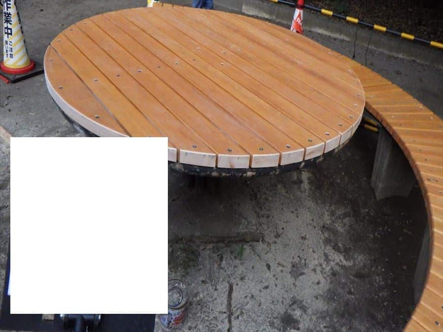 Circular wooden platform with slatted top and curved bench, outdoors at a construction site with cones in the background.