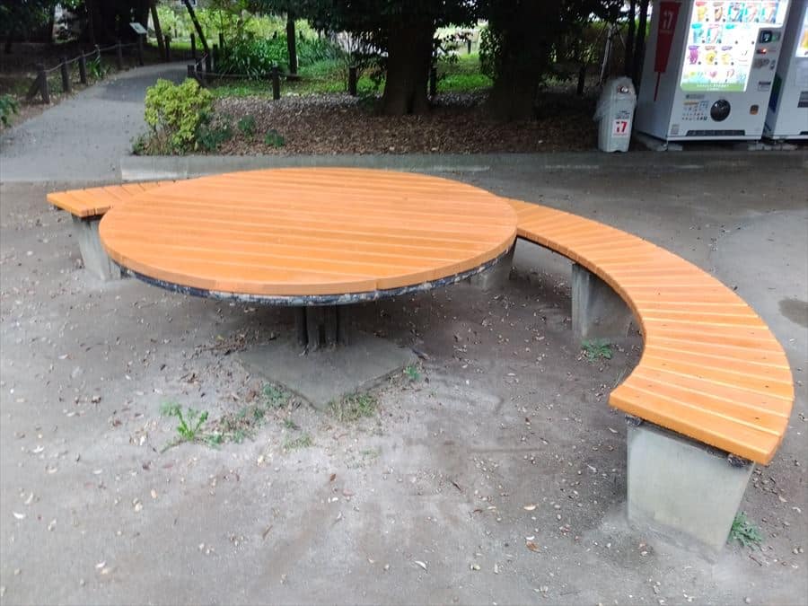 Circular orange wooden table with a curved surrounding bench in a park setting, under trees and near a vending machine area