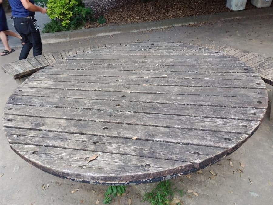 Large round weathered wooden outdoor table with benches, set on a concrete area in a park. A person’s legs are visible nearby.