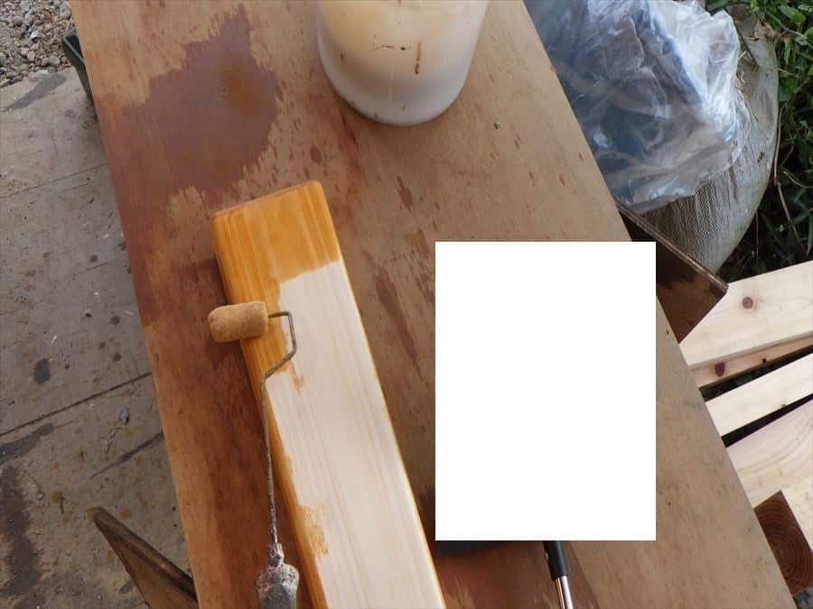Paint roller applying light varnish to a wooden plank on a worktable outdoors, with a bucket nearby.