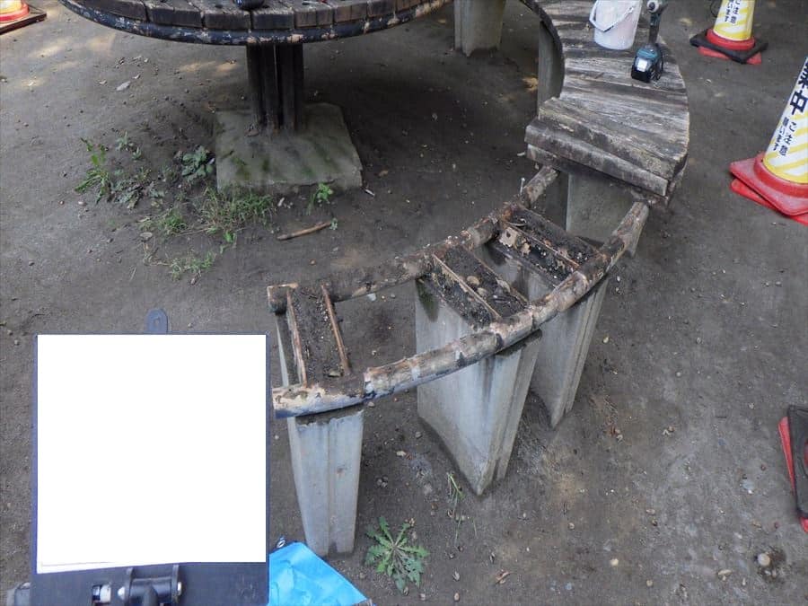 Worn, curved wooden bench frame with metal supports on dirt ground, under repair near construction cones in the background