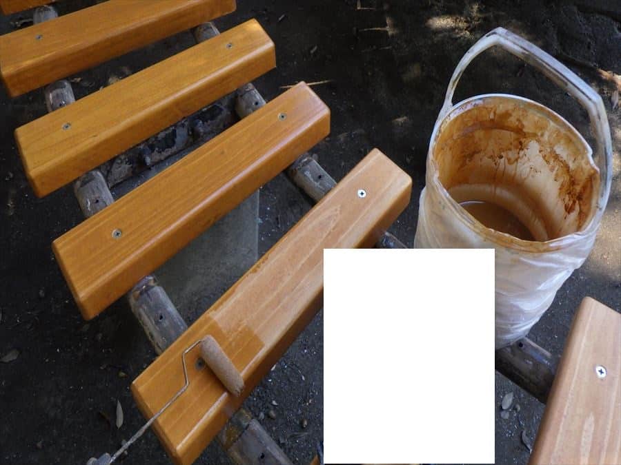 Outdoor wooden bench under maintenance with a paint roller resting on a slat and a paint bucket nearby, in a park setting.