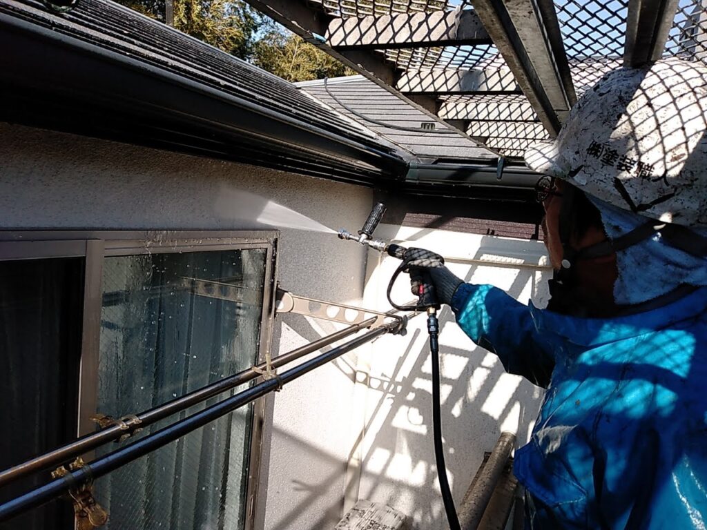 DCP PHOTO | 外壁塗装は神奈川県横浜市の塗装職人 Worker in blue protective gear and helmet power-sprays the exterior wall from a scaffold or balcony, cleaning the surface with a high-pressure nozzle.