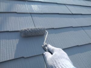 Close-up of a gloved hand using a paint roller to coat a blue-gray roof tile surface in spray-streaked light and shadow.