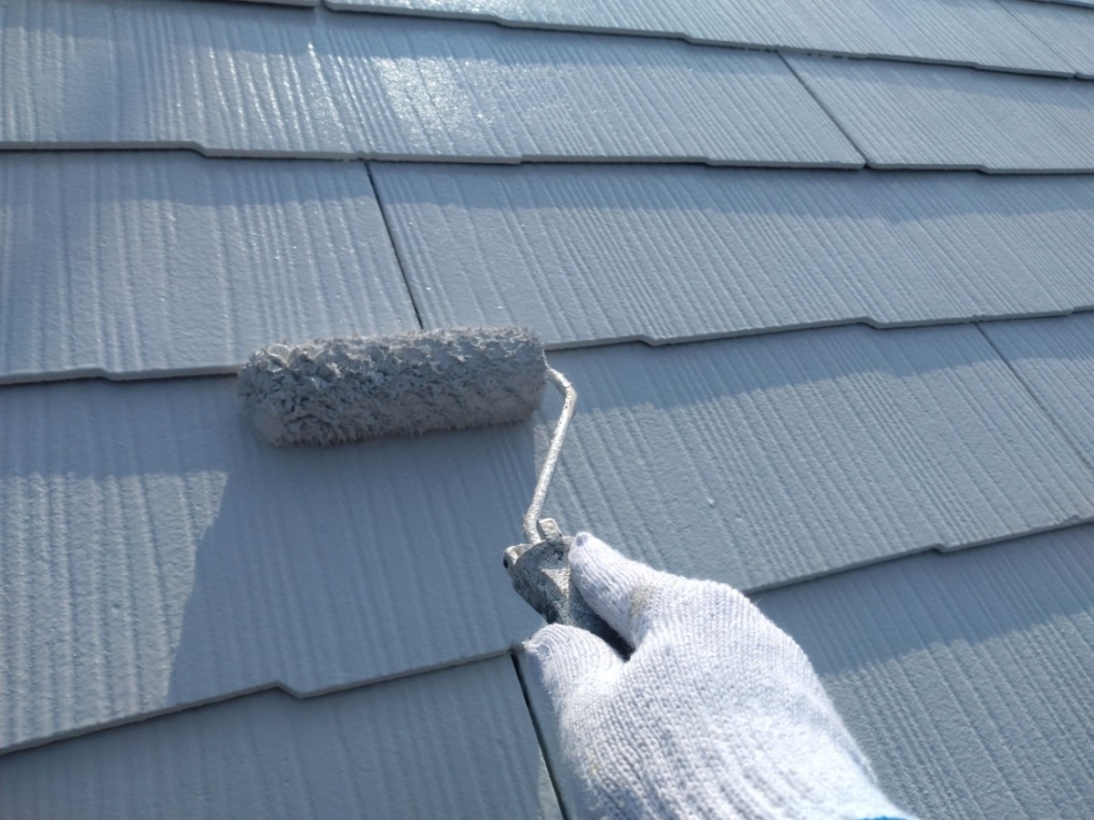 Close-up of a gloved hand using a paint roller to coat a blue-gray roof tile surface in spray-streaked light and shadow.