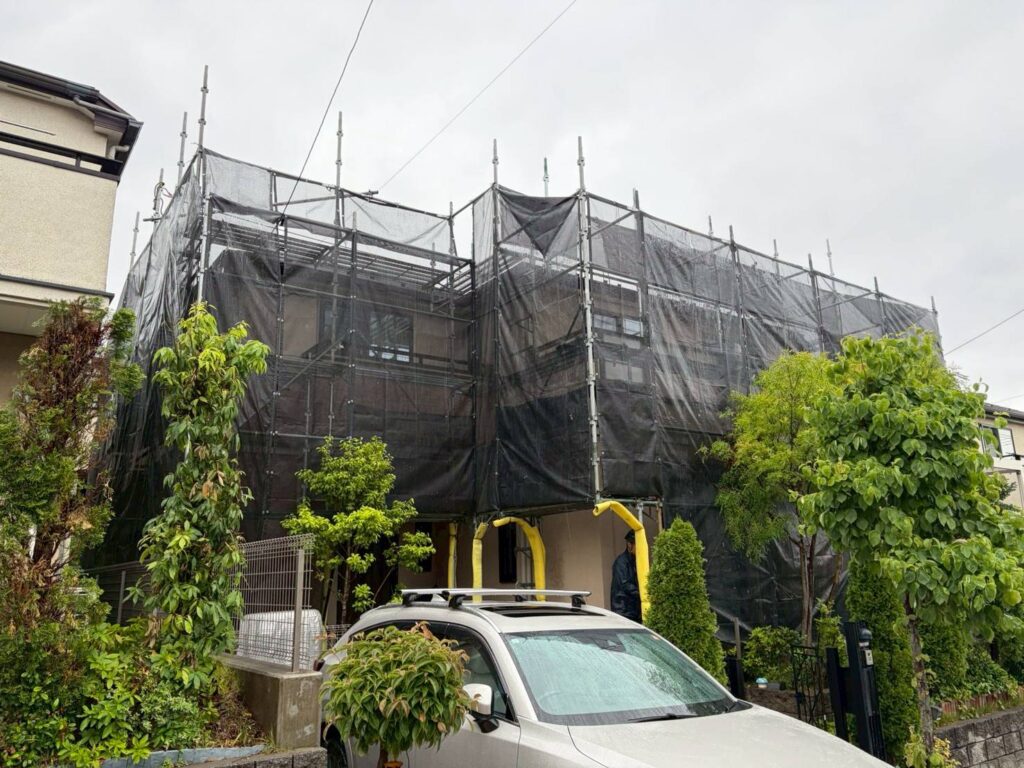 221010_0 | 外壁塗装は神奈川県横浜市の塗装職人 House under scaffolding with black netting covering during renovation; yellow safety arches at the entrance; car parked in front amid trees and shrubs.