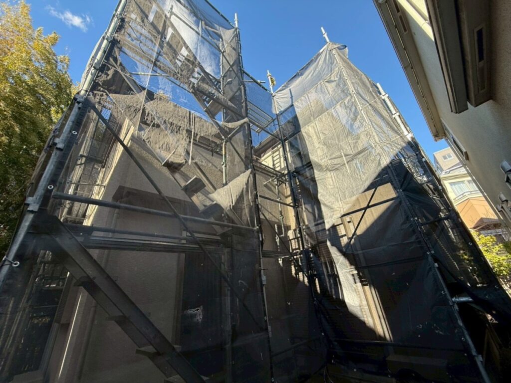 S__9404436 | 外壁塗装は神奈川県横浜市の塗装職人 Construction scaffolding with black safety mesh surrounding a residential building under renovation against a blue sky.