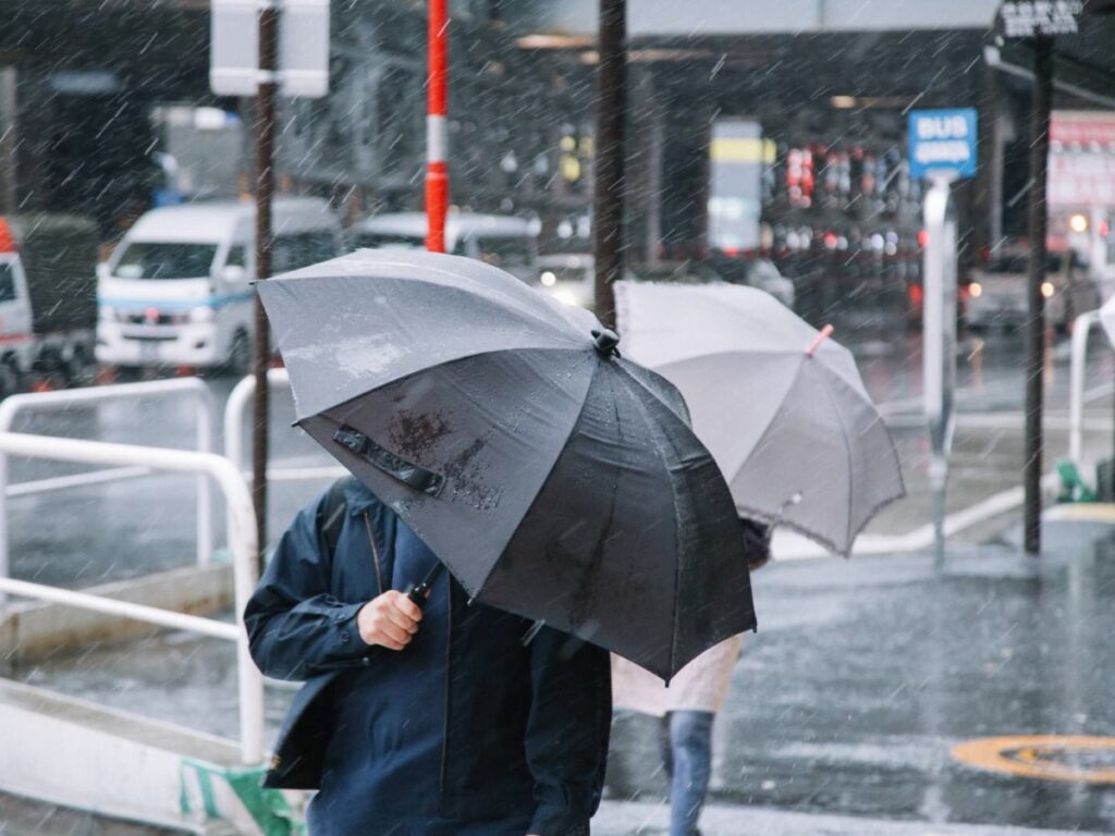 taihu-kasa0I9A5526_TP_V | 外壁塗装は神奈川県横浜市の塗装職人 Person holds a large black umbrella in a rainy urban street, with blurred city background and passing vehicles.