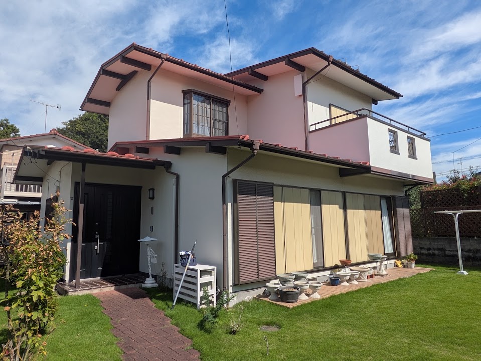 01-before | 外壁塗装は神奈川県横浜市の塗装職人 Two-story house with white walls, brown shutters, and a red-tiled roof, front lawn and row of potted plants along the patio doors under a blue sky.