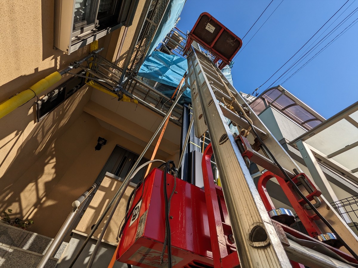 Construction setup outside a residential building: ladder and scaffolding reach upward with red equipment at ground level and overhead power lines against a blue sky