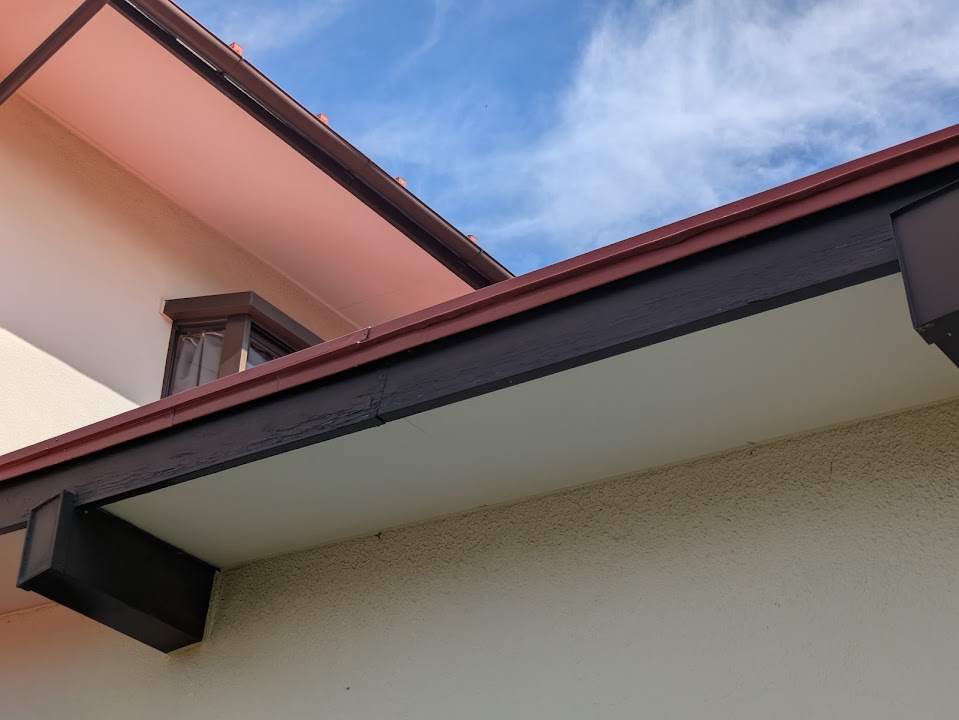 02-fascia-before | 外壁塗装は神奈川県横浜市の塗装職人 Close-up of a beige building with a dark brown wooden beam along a roofline and a blue sky above.